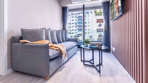 Ein modernes Wohnzimmer mit grauem Sofa, einem kleinen runden Tisch und einem großen Fenster mit Blick auf hohe Gebäude.