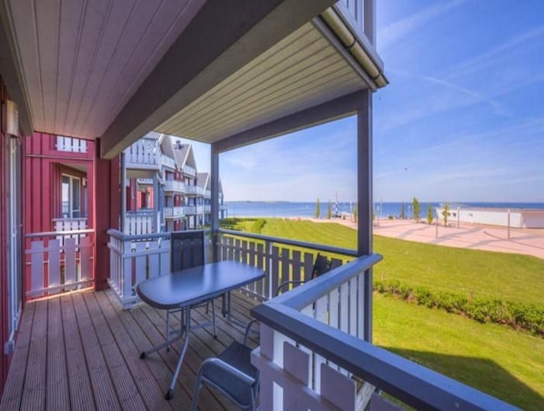 A wooden deck with a table and chairs, overlooking a beach and a building with red and white facades.