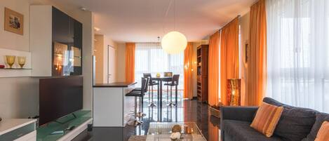 A modern living room with a sofa, coffee table, and a TV. There's a kitchen area with a dining table and chairs. The room has large windows with curtains.