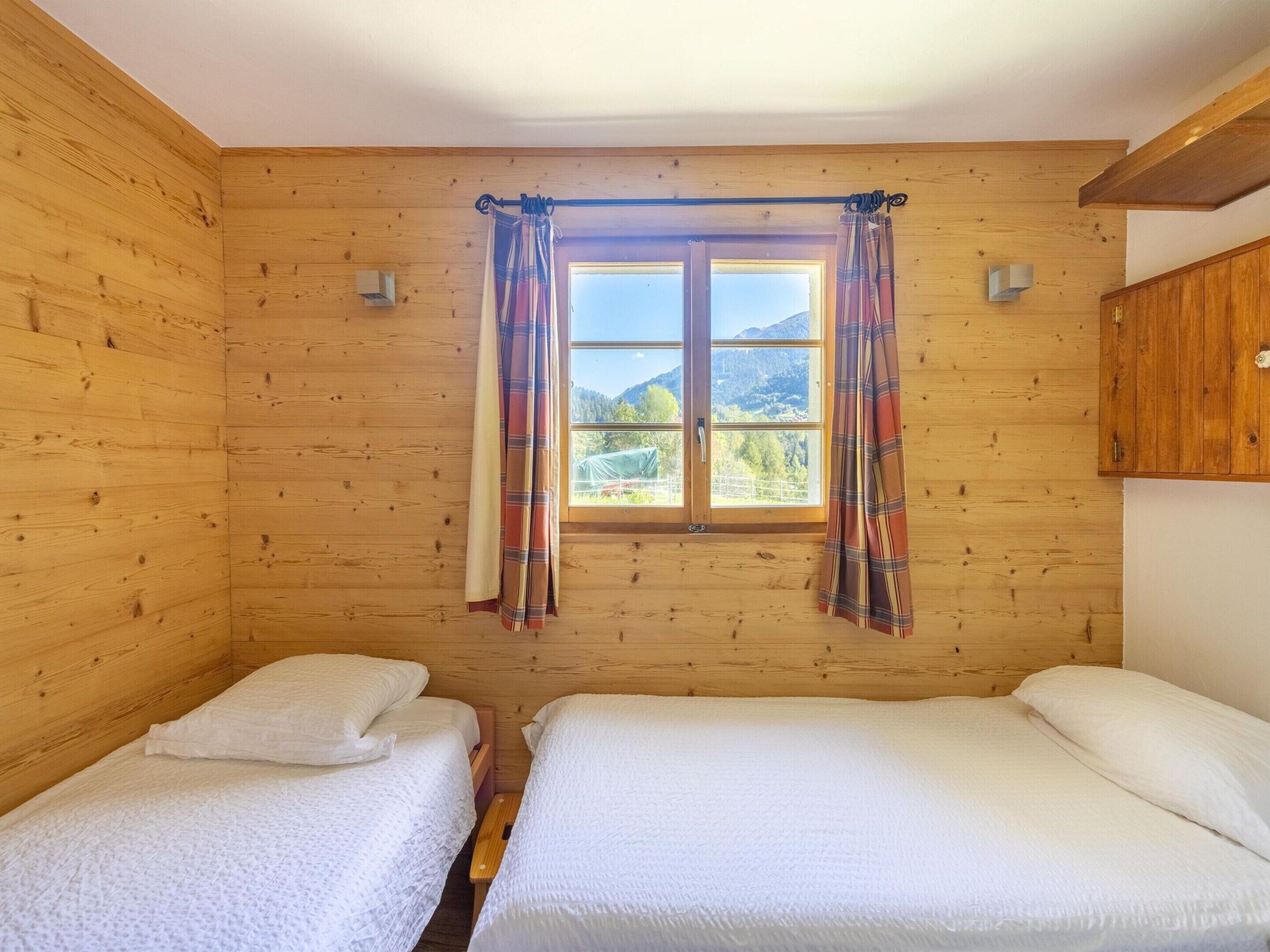 Une chambre dans un chalet en bois, avec deux lits, une fenêtre ornée de rideaux à carreaux et une vue sur les montagnes.