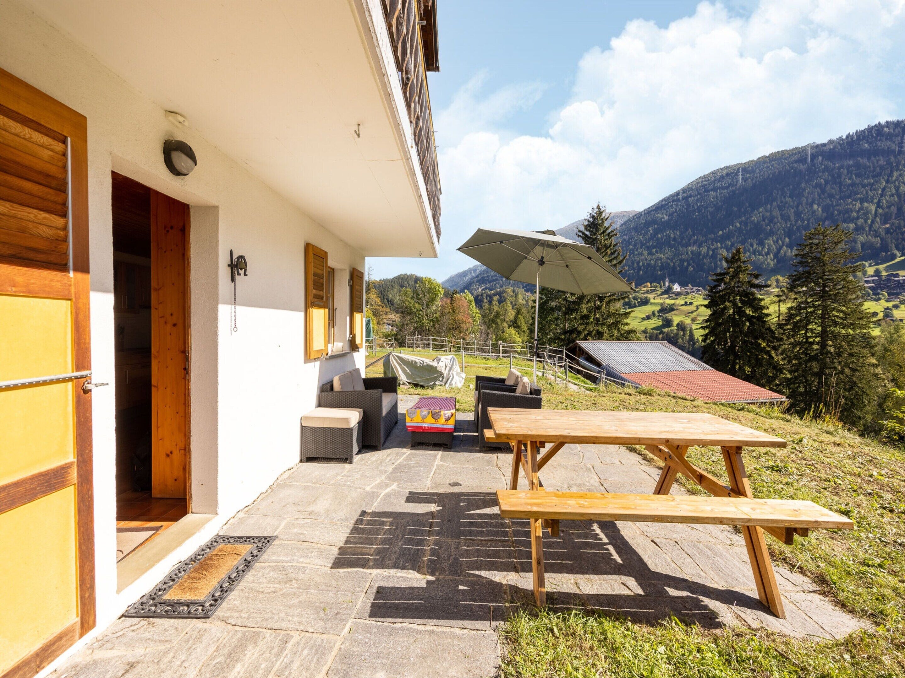 A patio with a wooden table and chairs, a covered area with a table and chairs, and a view of a mountainous landscape.