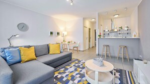 A modern living room with a grey sofa, yellow and blue cushions, a patterned rug, a round coffee table, and a kitchen area with stools and a bar. - Grand Place Central Gem 4

 (Bruxelles)