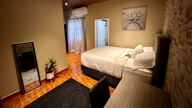 A bedroom with a bed, a desk, a chair, a potted plant, and a painting on the wall.