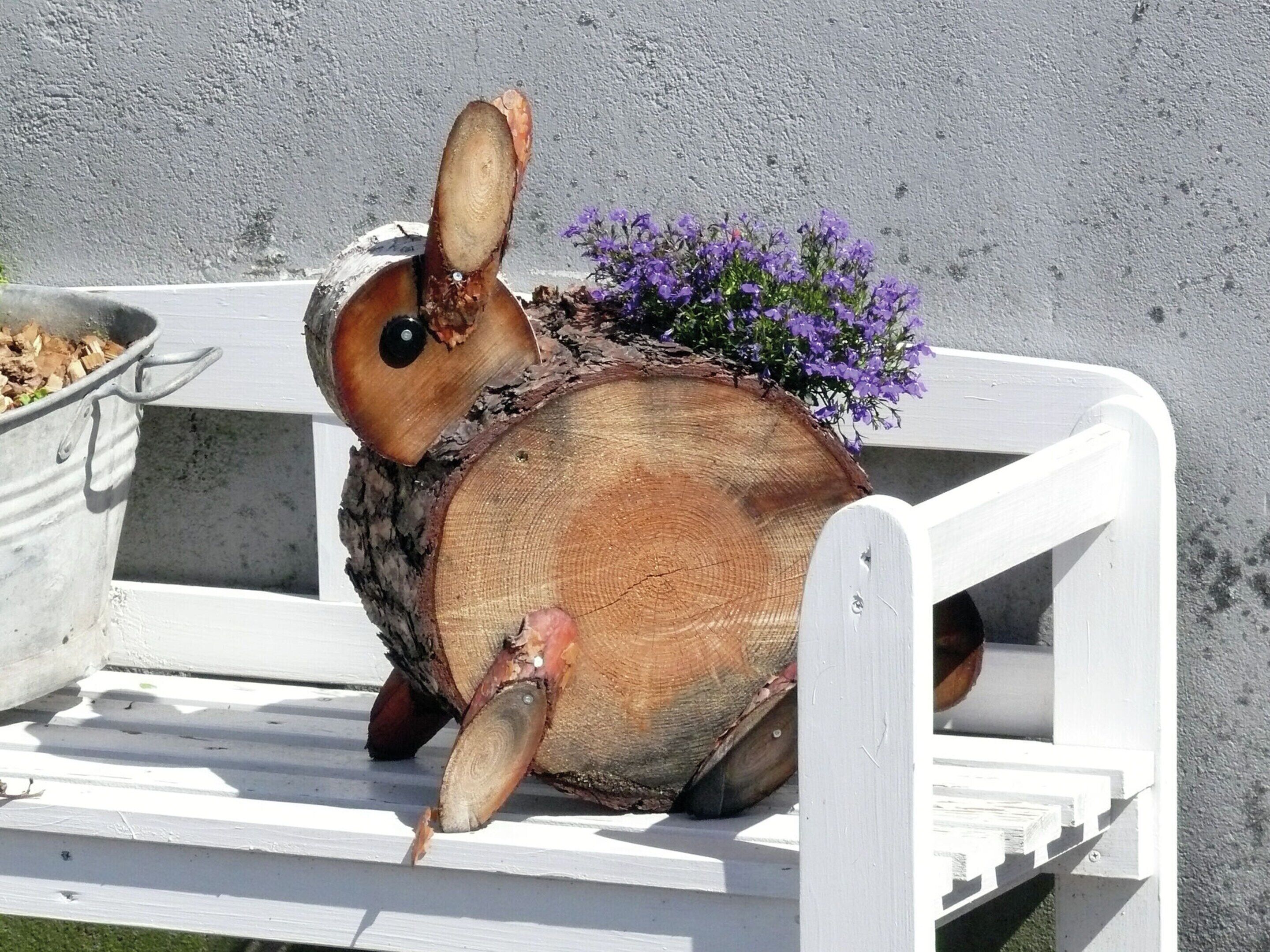 Una escultura de conejo de madera con cuerpo de tronco y orejas grandes, sobre un banco blanco frente a una pared con textura.