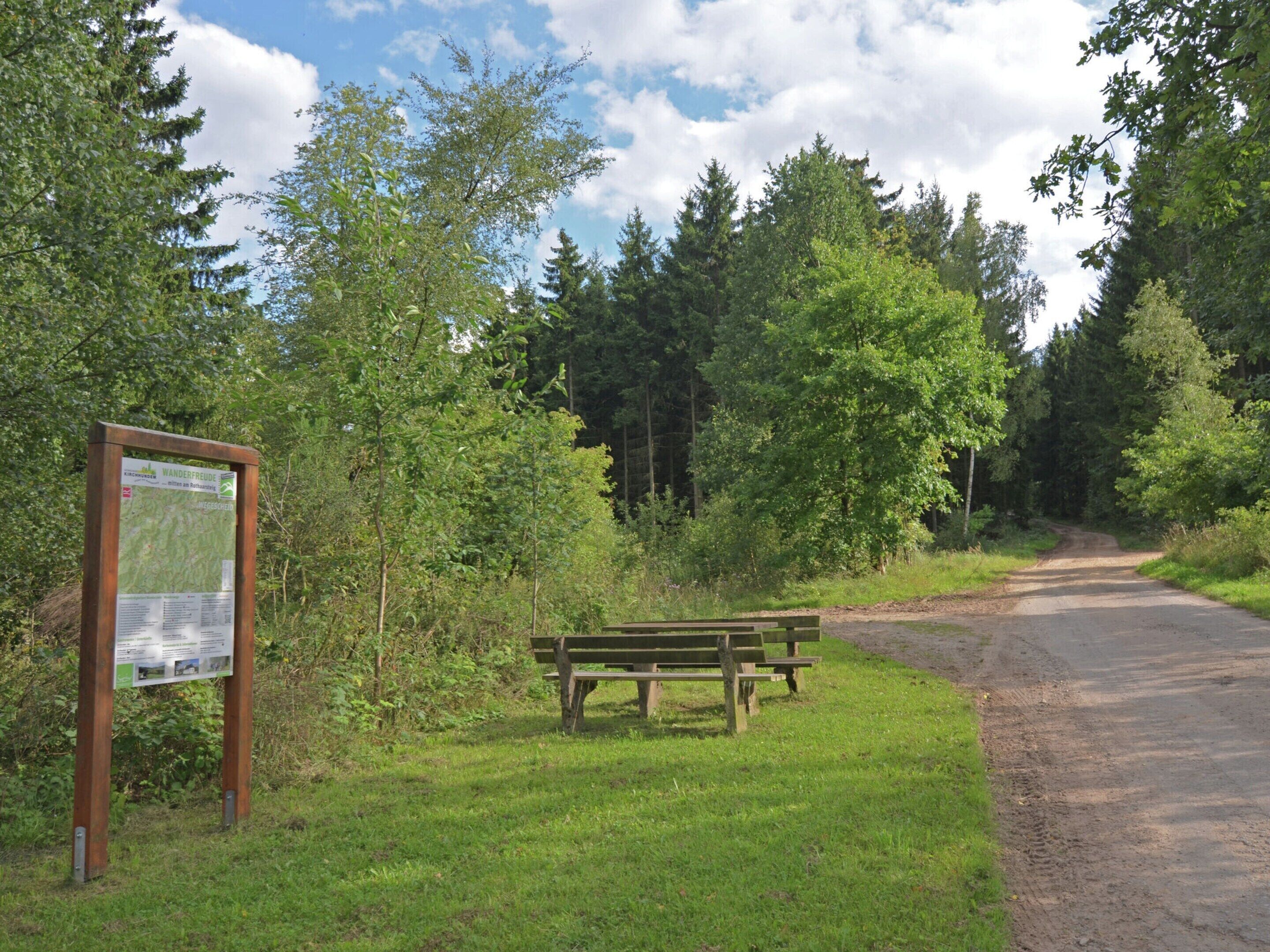 Un banc en bois et un panneau indicateur dans une zone boisée, le long d’un sentier de terre.