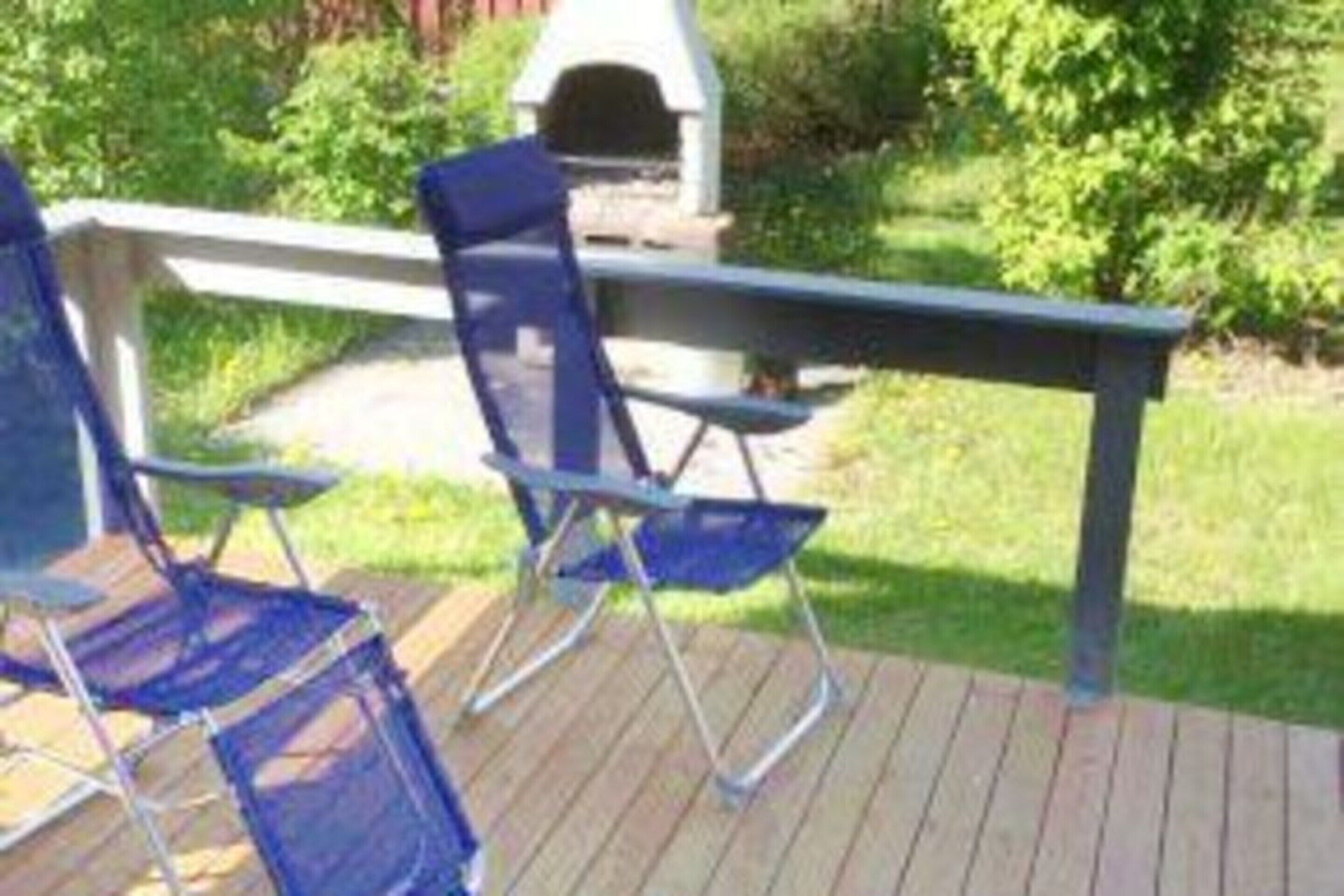 A deck with two blue folding chairs and a table, surrounded by greenery.