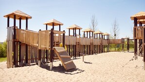 Eine hölzerne Spielanlage mit mehreren Ebenen und Rutschen auf sandigem Untergrund.