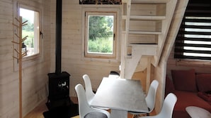 Interior acogedor de una cabaña con una mesa de comedor, sillas blancas, un hogar de leña negro y una zona superior con una escalera.