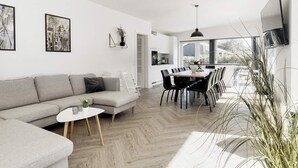 A modern living room with a grey sofa, a dining table with chairs, and a kitchen area visible through an open door. - 12 Person Holiday Home in Vaeggerlose (Vaeggerlose)