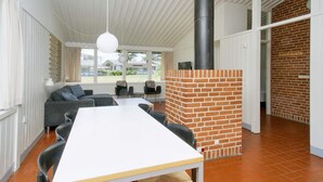 Et moderne interiør med hvidt loft, en murstenspejs, et spiseområde med bord og stole, og udsigt til det fri gennem store vinduer.