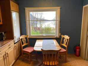 A small kitchen with wooden cabinets, a microwave, a window with a view of trees and a house, and a table with two chairs. - 6 Person Holiday Home in Storslett (Nordreisa)