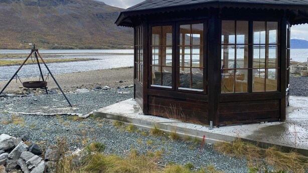A wooden gazebo with a sloped roof overlooking a lake, surrounded by rocks and sparse vegetation. - 6 Person Holiday Home in Storslett (Nordreisa)