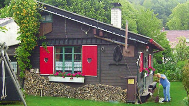 A rustic wooden house with red shutters, a chimney, and a garden area with a person tending to plants. - Ferienhaus in Wieda mit Garten (Walkenried)