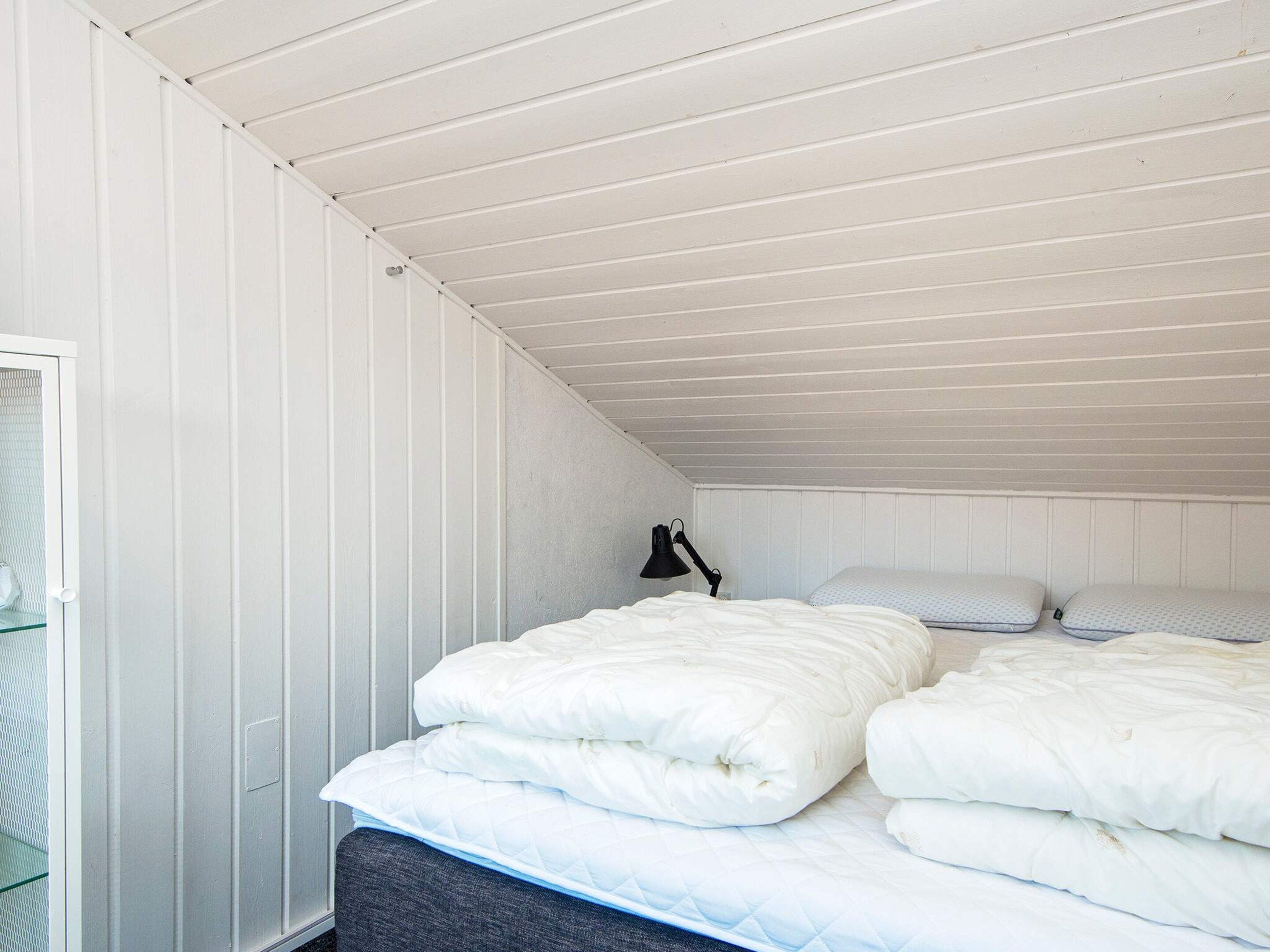 Ein Schlafzimmer mit schrägemausertem Dach, einem Bett mit weißer Bettwäsche, einer Nachttischlampe und einem Fenster mit Jalousien.