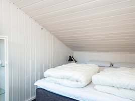 Un dormitorio con techo inclinado, una cama con ropa de cama blanca, una lámpara de noche y una ventana con persianas.