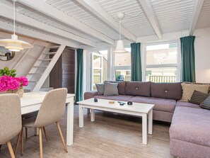 Una sala de estar moderna con un sofá, una mesa de comedor con sillas, una escalera y ventanas grandes con cortinas.