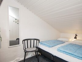 Un dormitorio con techo inclinado, una cama individual con ropa de cama azul, una silla con cojín y una mesita de noche con lámpara.