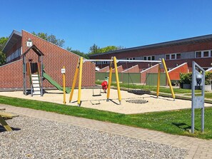 Ein Spielplatz mit Schaukeln und Rutsche, im Hintergrund ein Backsteinhaus und ein Kiesweg.