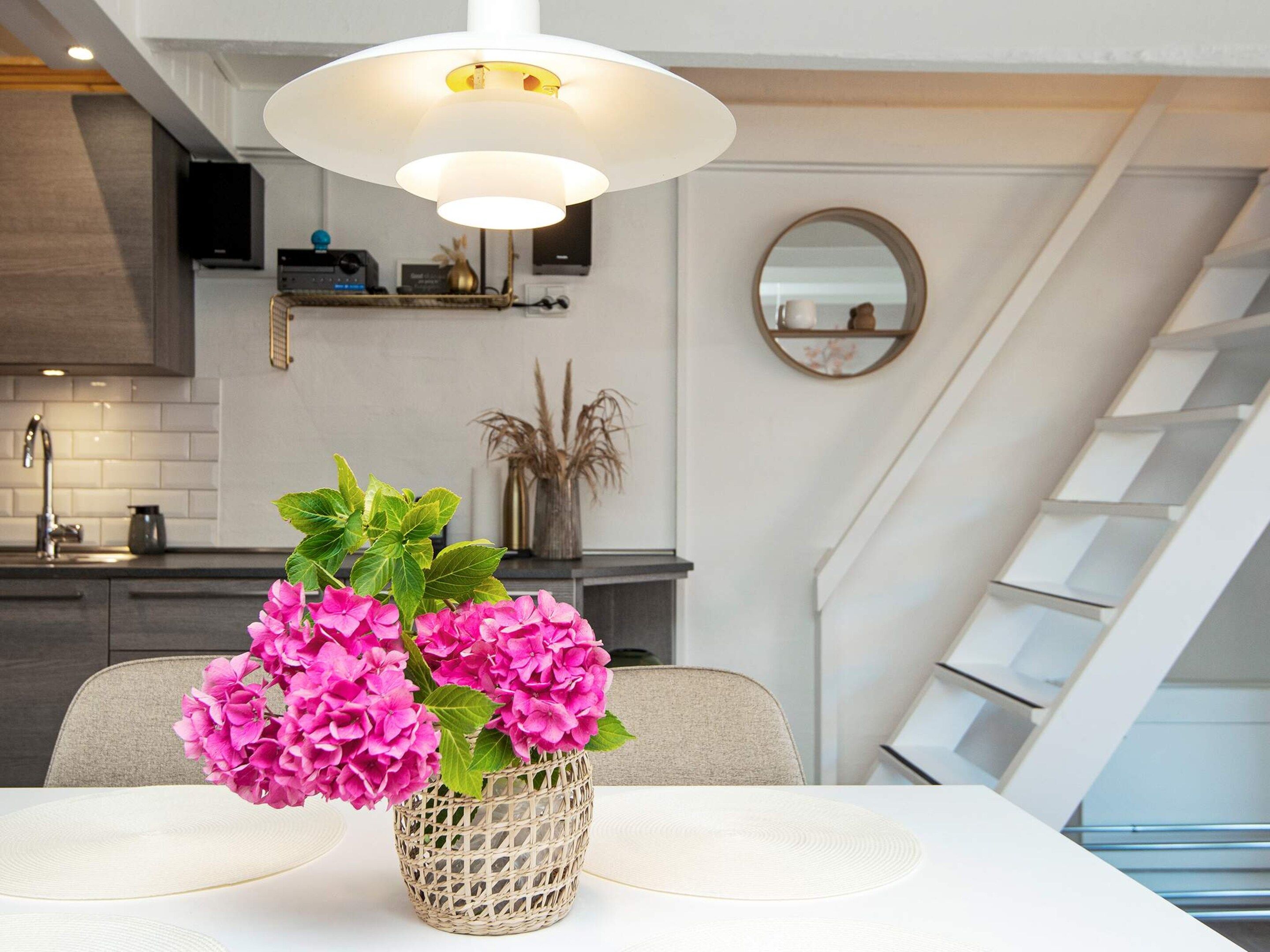 Een moderne keuken met een witte plafondlamp, een eettafel met een gevlochten mand vol roze bloemen en een trap met een witte leuning.