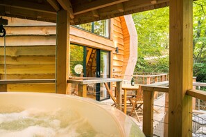 Eine Holz-Blockhütte mit Whirlpool, einer Holzdiele und Blick auf den umliegenden Wald.