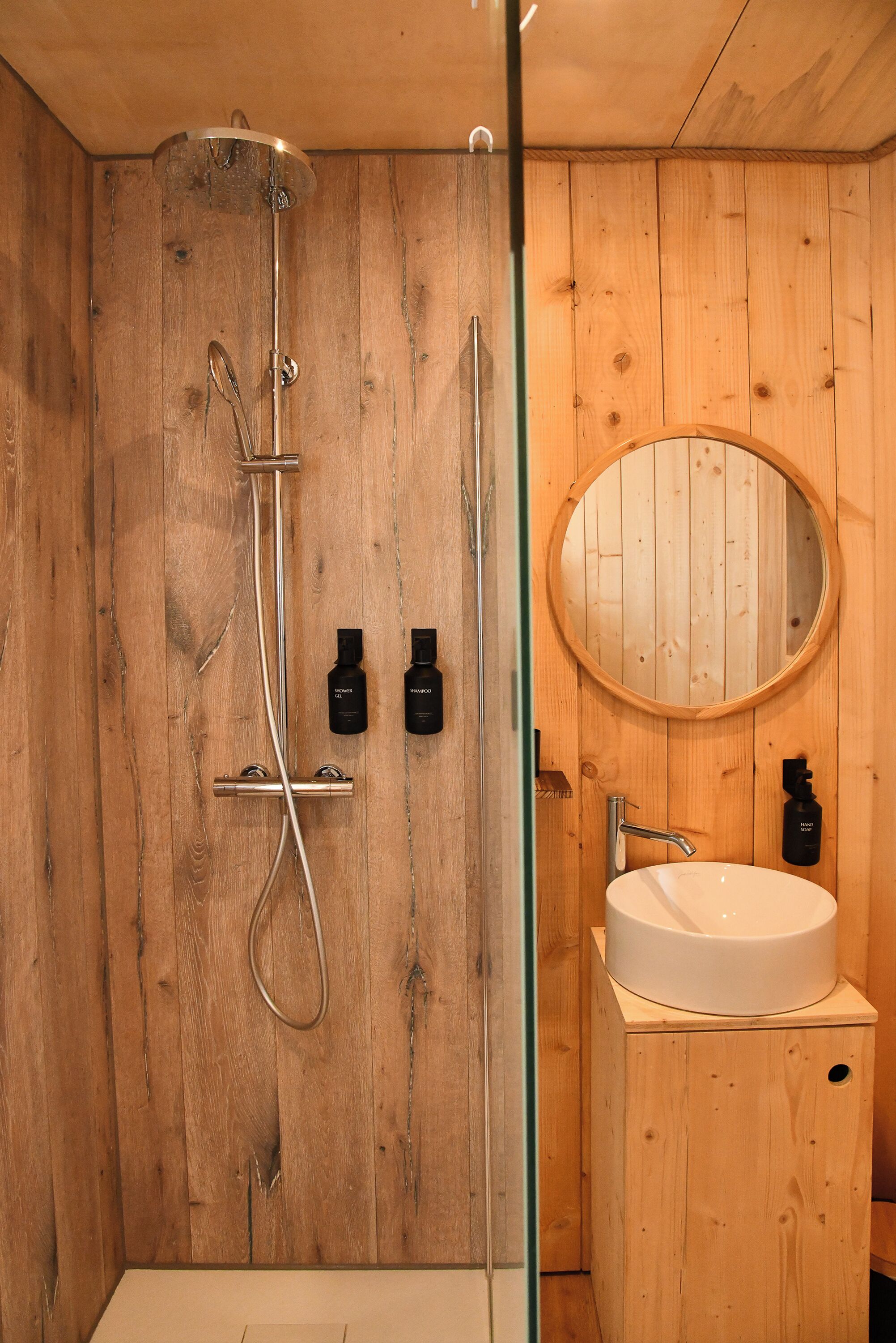 A wooden-paneled bathroom with a shower, a sink, and a round mirror.