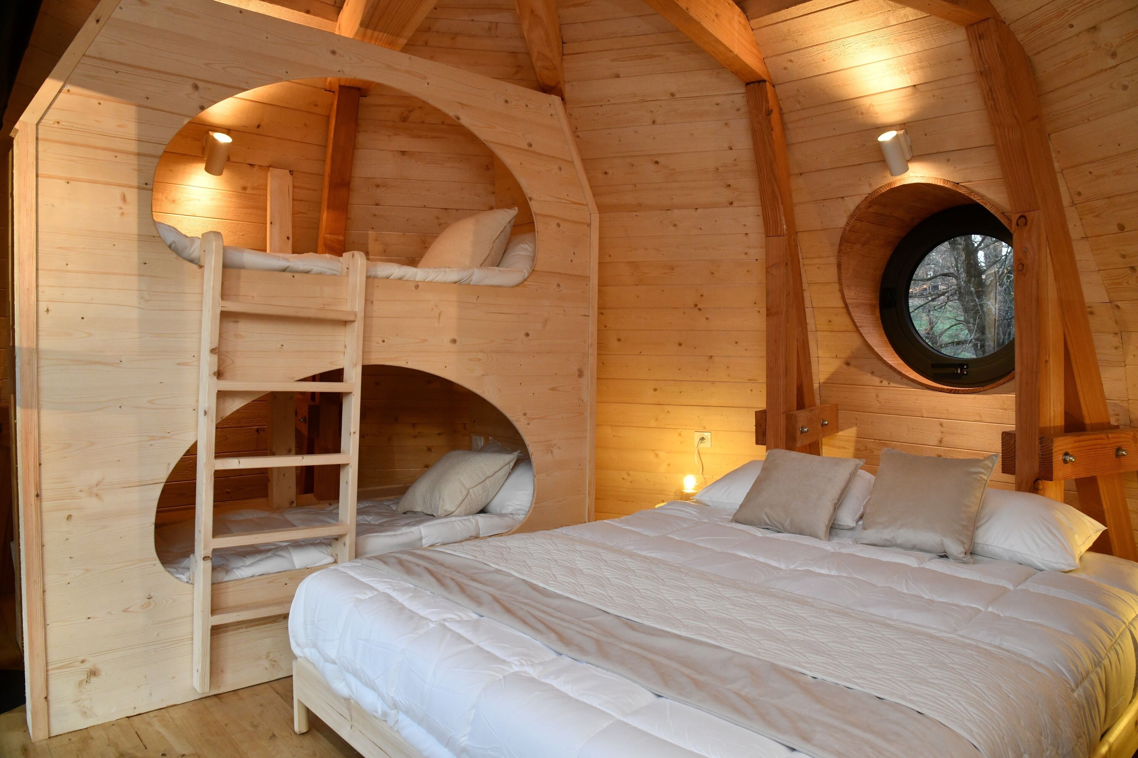 Une cabane en bois avec un lit superposé, une fenêtre ronde et une échelle.