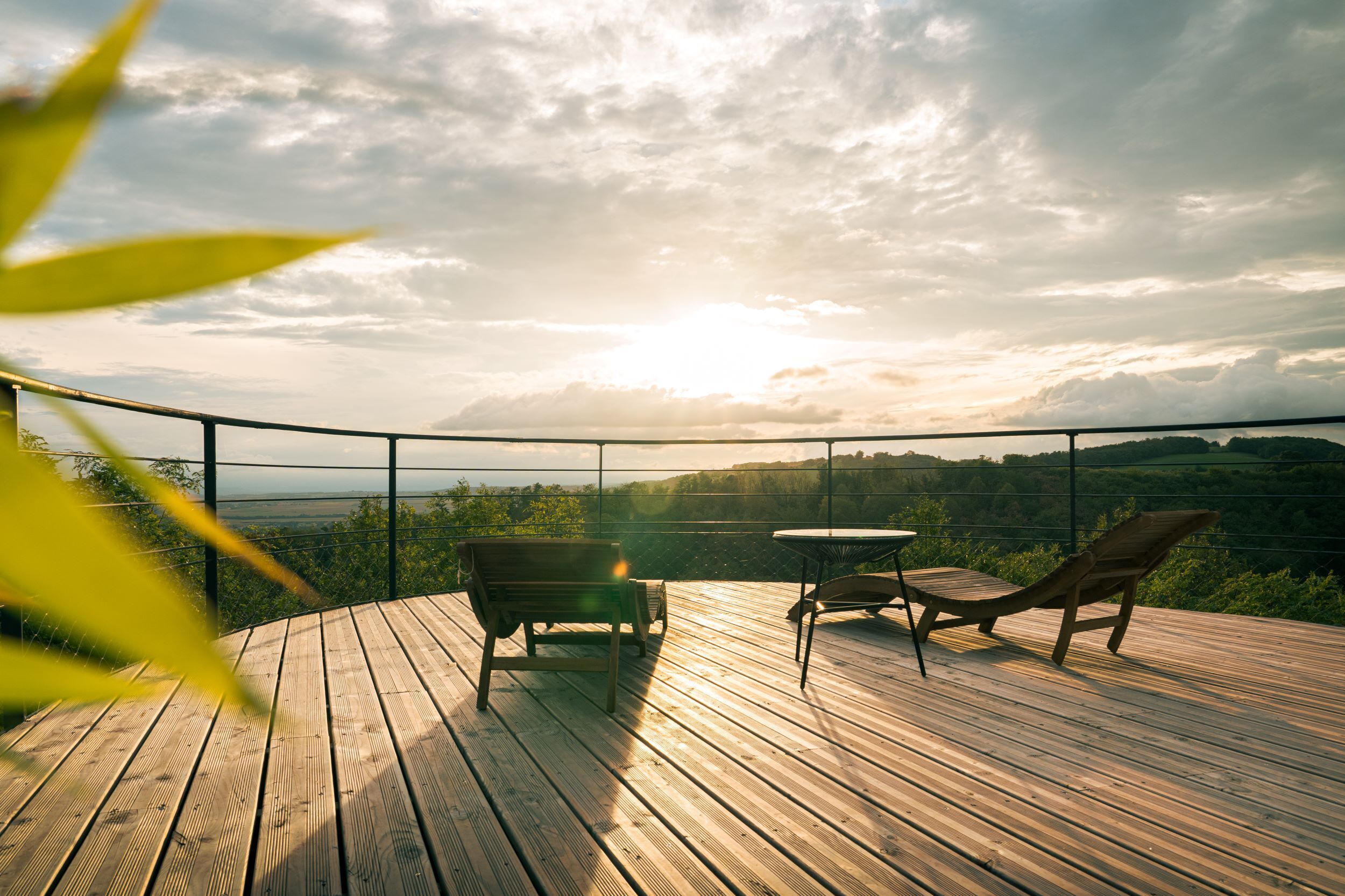 A wooden deck with two lounge chairs and a table, overlooking a scenic landscape.