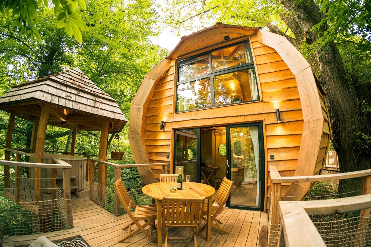 Uma casa na árvore de madeira com um deck, uma área de refeições com cadeiras e mesa, e uma porta de vidro que dá para o interior.