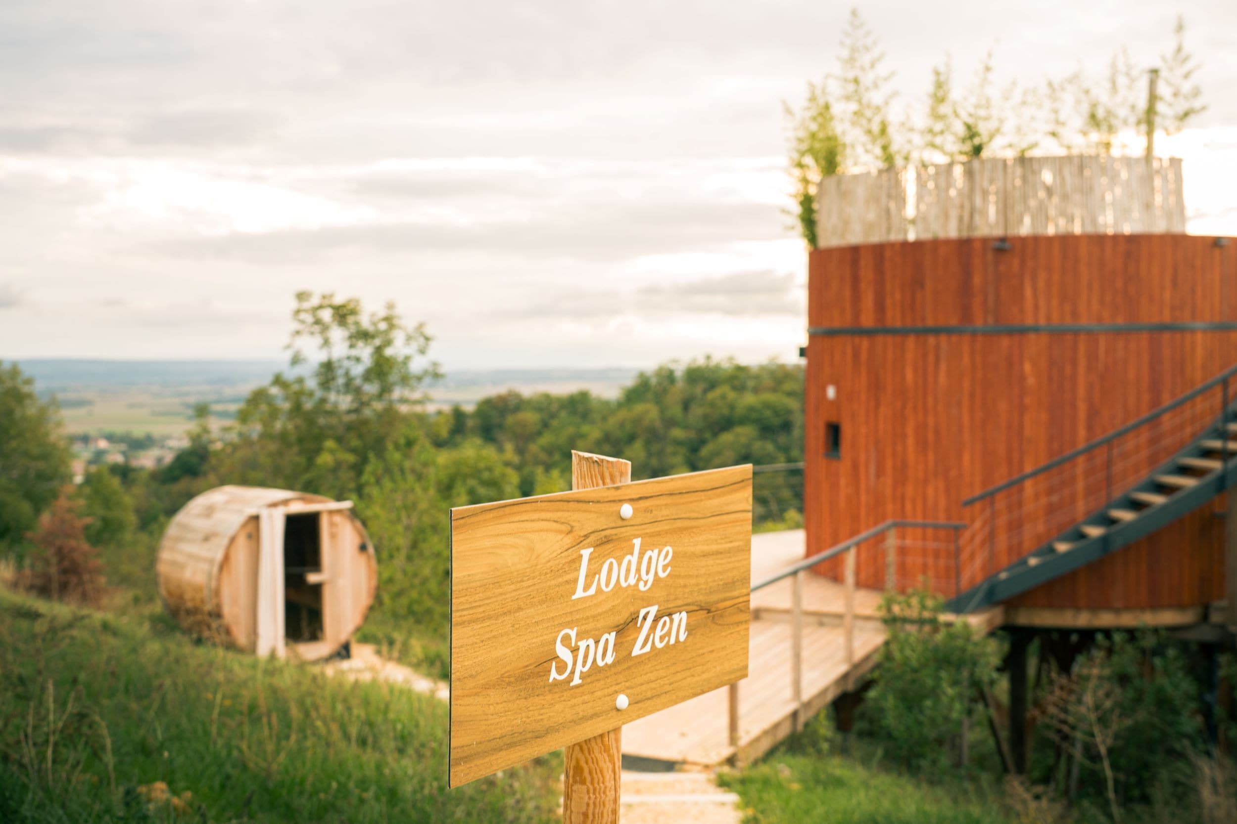 A wooden sign reading "Lodge Spa Zen" with a wooden barrel and a wooden structure in the background.