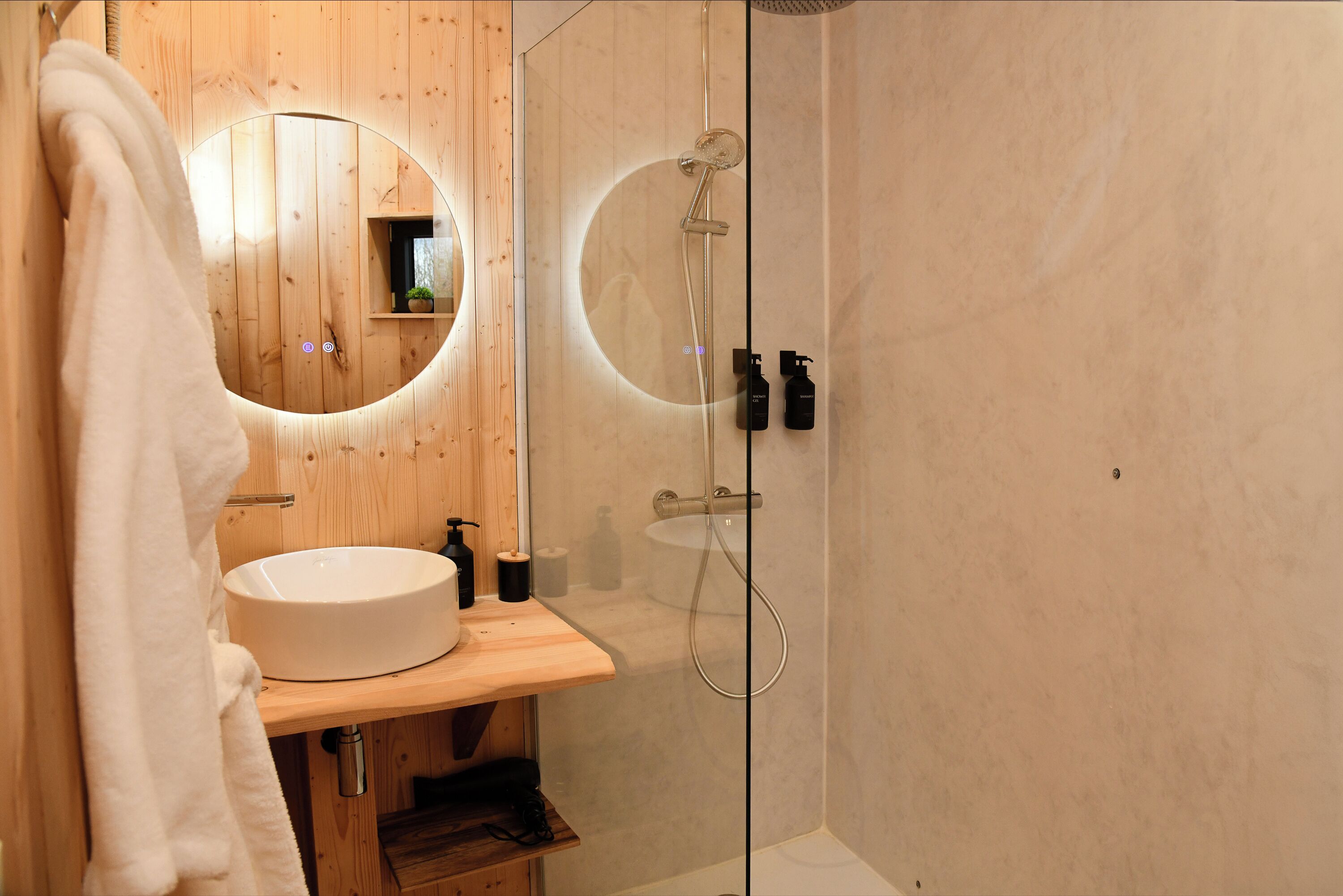 A bathroom with a wooden vanity, a round mirror, a sink, and a glass-enclosed shower area.
