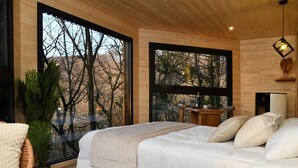 Una camera da letto con un letto grande, una pianta in vaso e vista su alberi attraverso ampie finestre.
