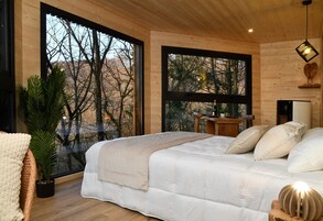 A bedroom with a large bed, a potted plant, and a view of trees through large windows.