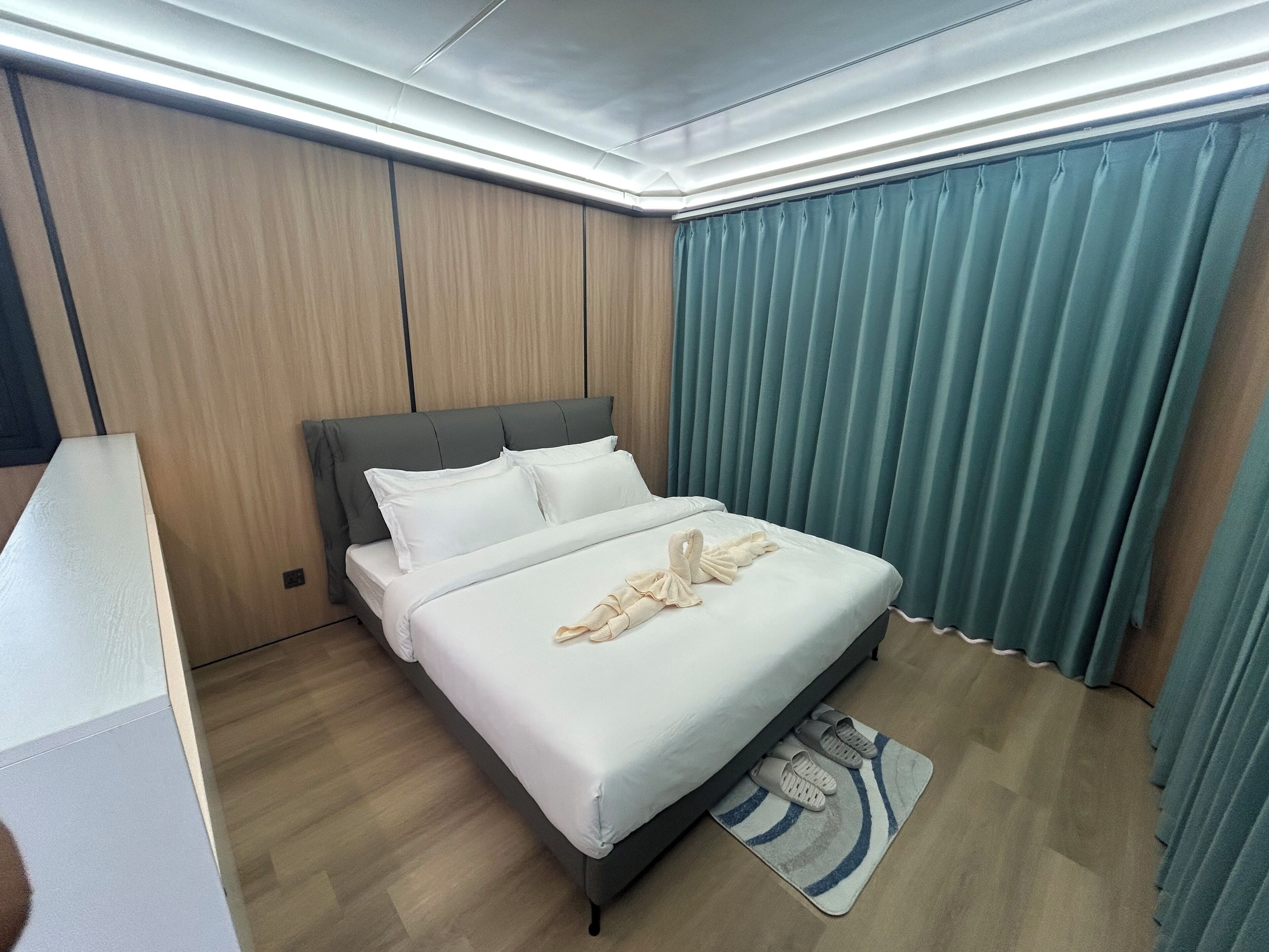 A modern hotel room with a large bed, wooden panel walls, and a view of the city through floor-to-ceiling windows.