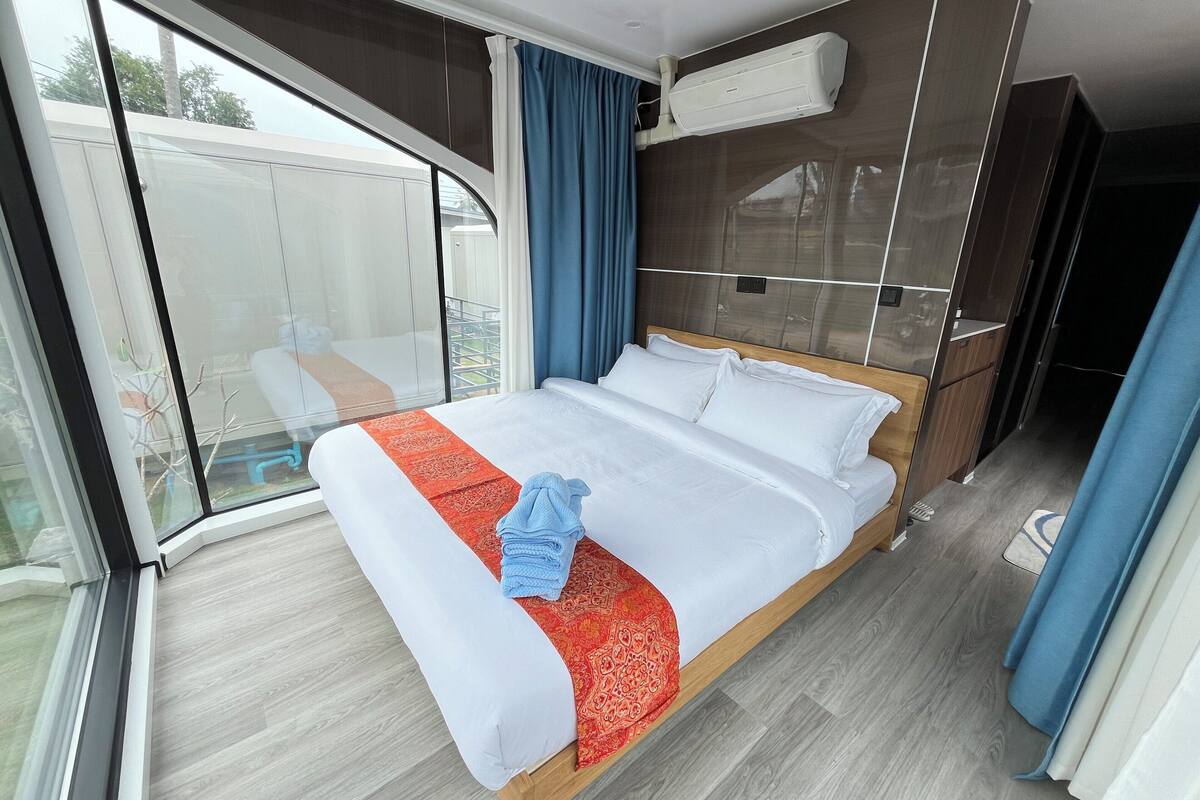 A hotel room with a large bed, a window with a view, a ceiling air conditioner, and a wooden floor.