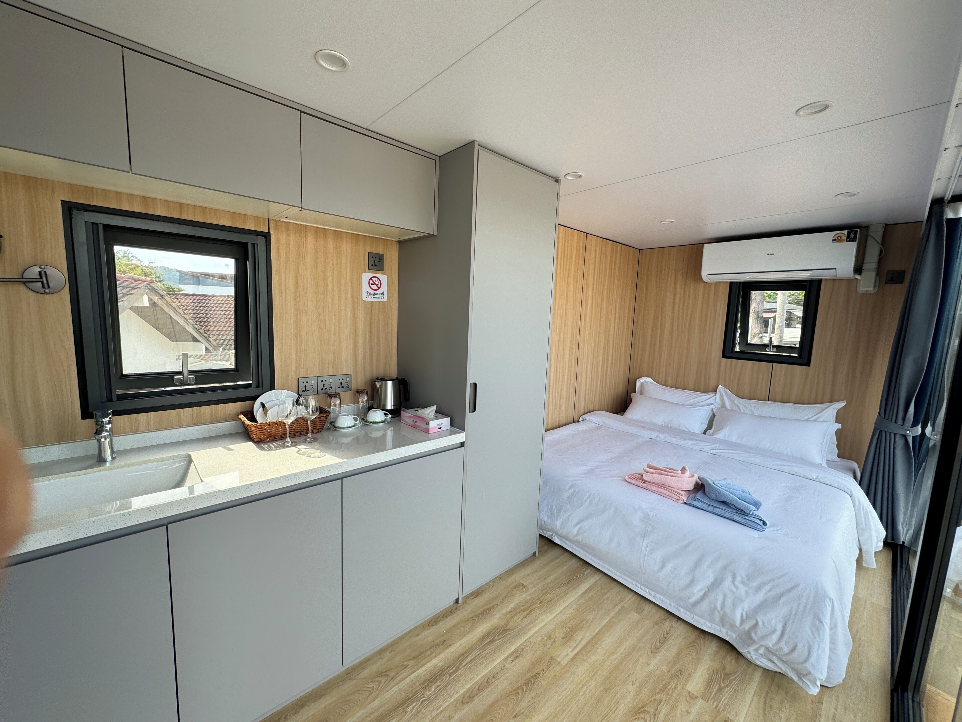 A compact bedroom with a bed, a small kitchenette, a window, and an air conditioning unit.