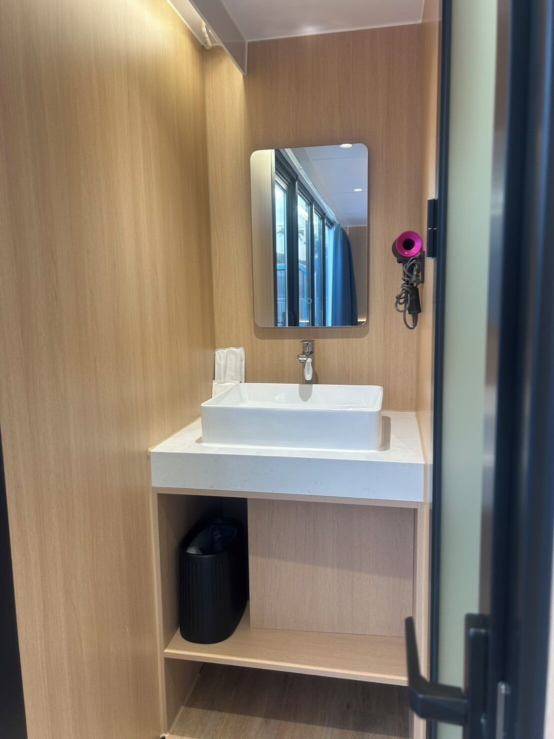 A modern bathroom with a white sink, a mirror, and a black trash bin.