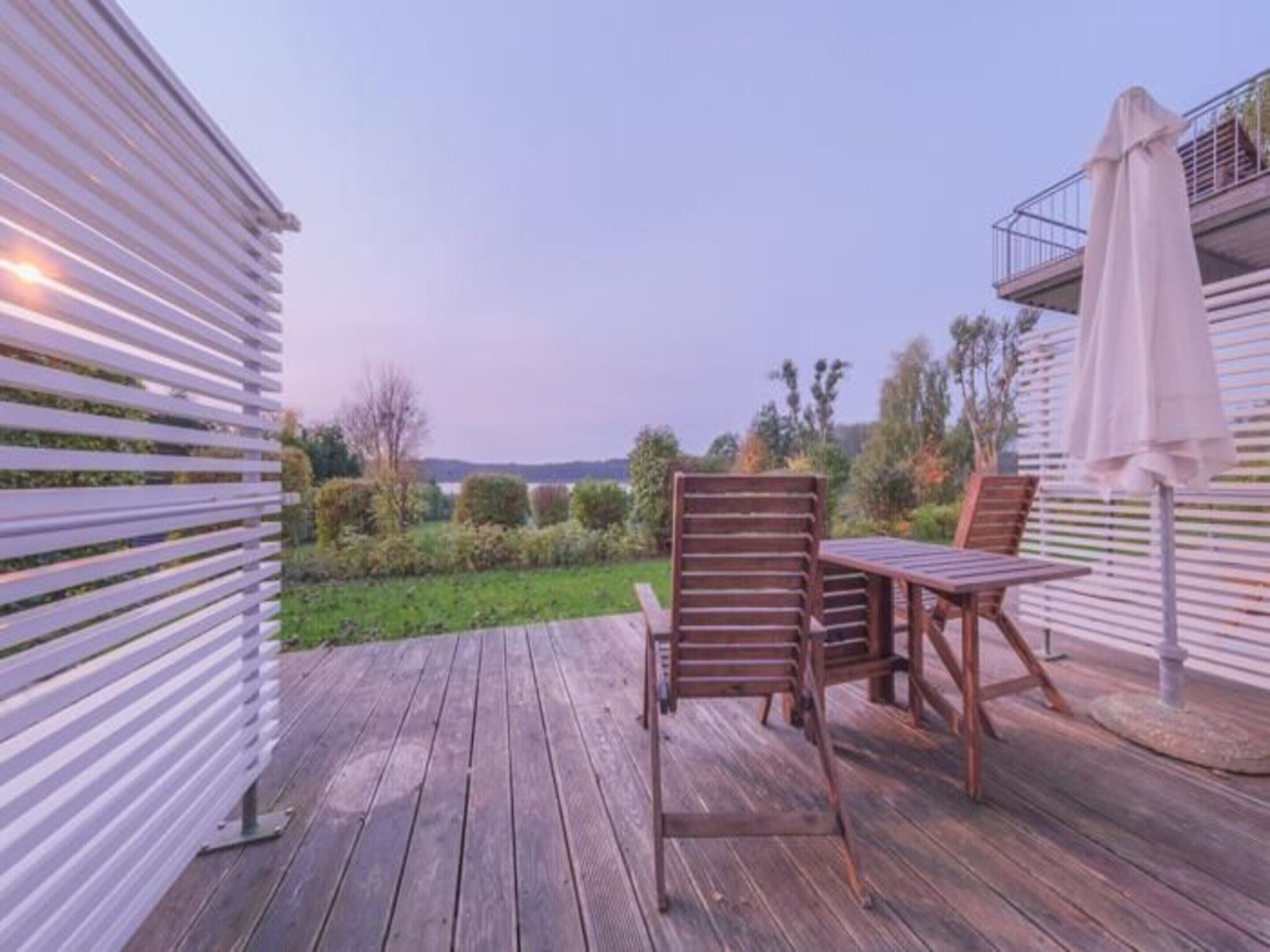 Um deck de madeira com duas cadeiras e uma mesa, cercado por uma cerca branca e vista para a paisagem.