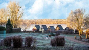 A row of traditional wooden cabins with sloped roofs, a grassy area with bushes, and a lake in the background. - Gemutliches Appartement mit Skandinavischem Flair (Mirow)