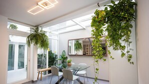 A modern dining area with a glass table and chairs, surrounded by large windows and indoor plants. - Villa Naim Guest House Cala del Moral (Rincon de la Victoria)