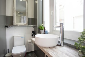 Een badkamer met een wit toilet, een wastafel met een witte kom, een spiegel boven de wastafel en een raam voor natuurlijk licht.