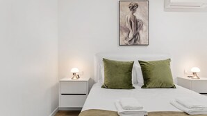 A modern hotel room with a bed, two nightstands, a wall-mounted air conditioner, and a framed artwork above the bed.