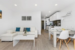 A modern living room with a white sofa, a coffee table, and a dining area with a table set for four. - Serene Blue Escape by Madeira Sun Travel (Ponta do Sol)
