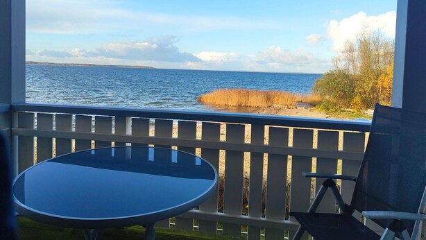 A balcony with a table and chairs, overlooking a body of water and a grassy area. - Appartement im Ferienpark in Rechlin (Rechlin)