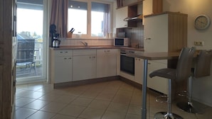 A compact kitchen with a dining area, a window with curtains, and a wall clock. - Appartement im Ferienpark in Rechlin (Rechlin)