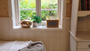 A bedroom with a window, a bed, a bedside table with a lamp, a small speaker, and a bookshelf.