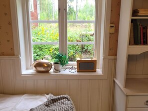 Quarto com janela, cama, mesa de cabeceira com abajur, um pequeno alto-falante e estante de livros.