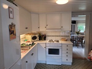 Une cuisine compacte avec des meubles blancs, un four à micro-ondes, un four traditionnel et un évier. On aperçoit à l’arrière-plan un coin repas avec une table et des chaises.