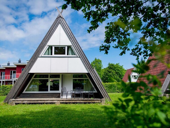A modern, triangular-shaped building with a flat roof, large windows, and a small patio area with outdoor seating. - Ferienhaus im Ostseeresort Dampland (Damp)