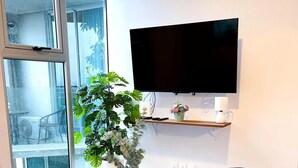 Een woonkamer met een bank, een salontafel met een bloemenvaas, een flatscreen televisie aan de muur en een klein blauw poef.