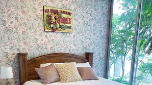 Een slaapkamer met bloemenbehang, een houten hoofdbord en een groot raam met uitzicht op bomen.